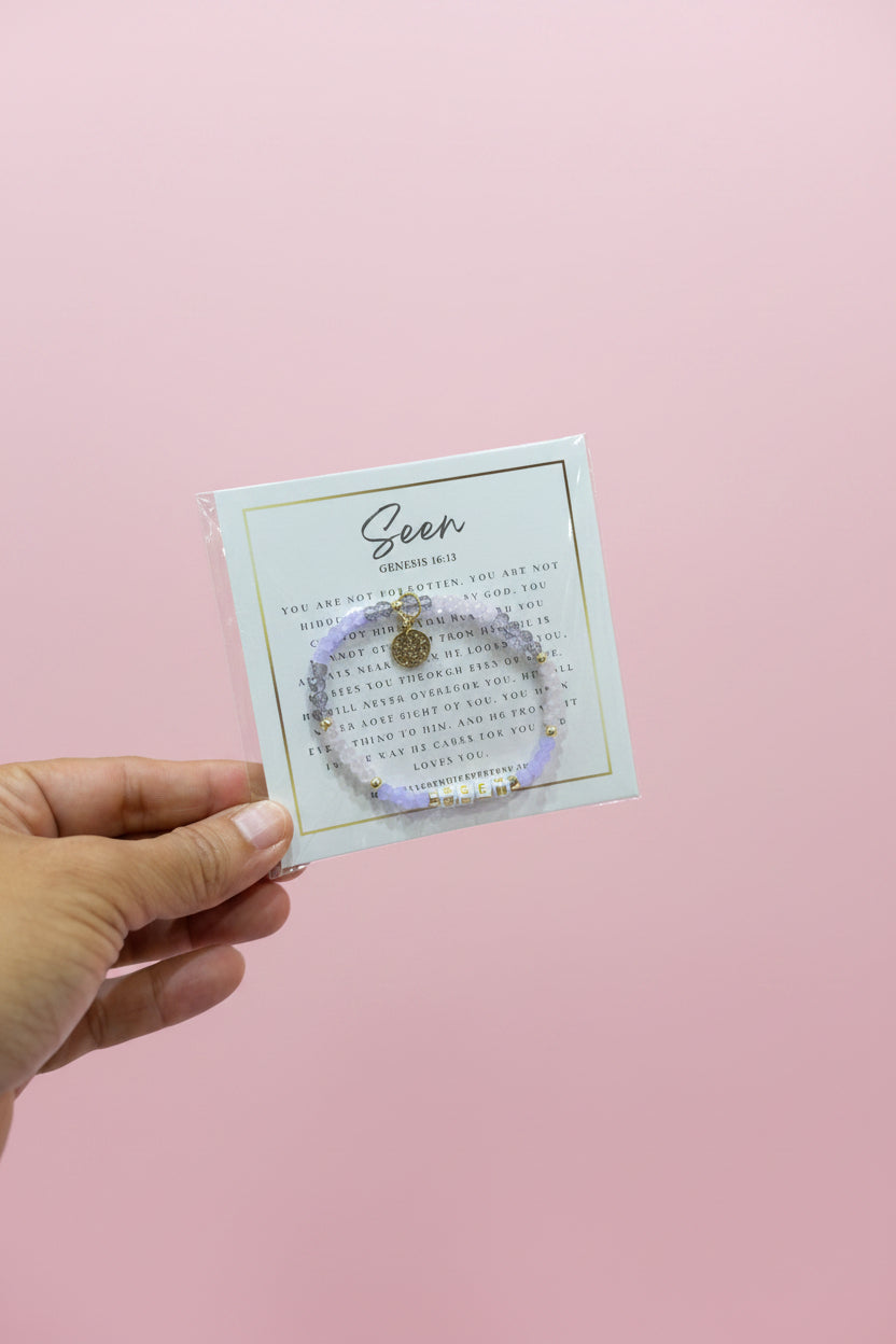 A hand holds a small bracelet on a display card in a jewelry store. The bracelet is light purple with a small gold charm and clear rhinestone accents.