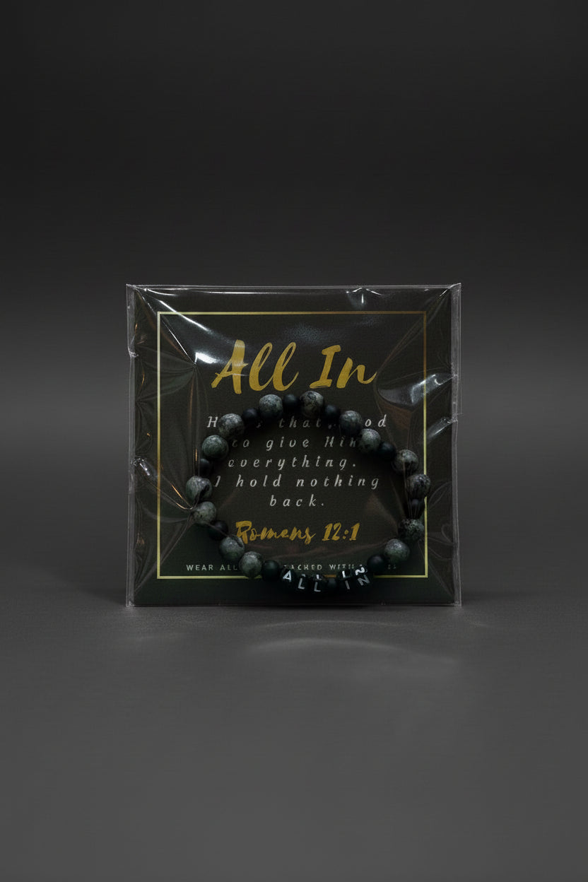 A hand holding a packaged beaded bracelet with a black backing card that reads 'All In' and a Bible verse from Romans 12:1. The bracelet itself appears dark gray to black with marbled stone-like texture.