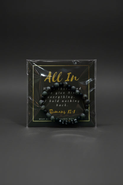 A hand holding a packaged beaded bracelet with a black backing card that reads 'All In' and a Bible verse from Romans 12:1. The bracelet itself appears dark gray to black with marbled stone-like texture.