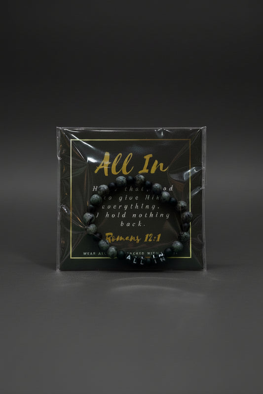 A hand holding a packaged beaded bracelet with a black backing card that reads 'All In' and a Bible verse from Romans 12:1. The bracelet itself appears dark gray to black with marbled stone-like texture.