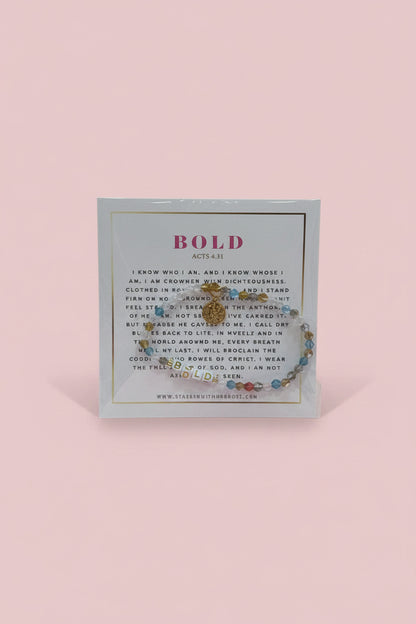 A hand holds a display card featuring a beaded bracelet with a gold-toned circular charm. The card has the word 'BOLD' printed at the top and a small brand name at the bottom.