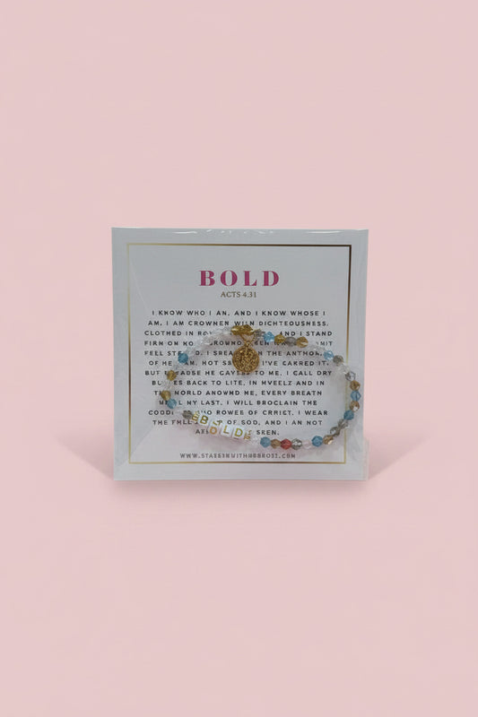 A hand holds a display card featuring a beaded bracelet with a gold-toned circular charm. The card has the word 'BOLD' printed at the top and a small brand name at the bottom.