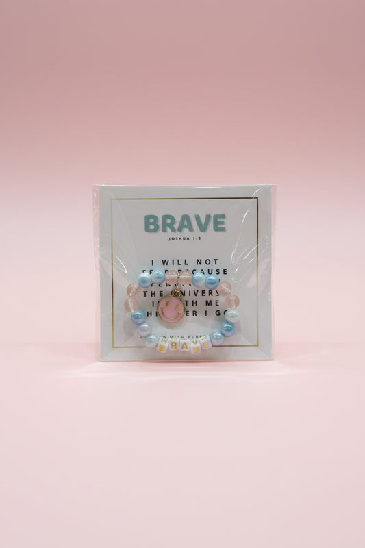 A hand holds a packaged beaded bracelet card in front of a display of similar cards. The front card reads "BRAVE" and shows a pastel beaded bracelet with a smiley face charm and letter tiles spelling BRAVE.