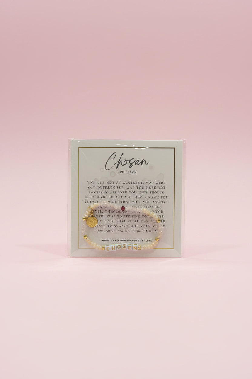 A hand holds a small gold beaded bracelet on a branded display card labeled 'Chosen Letters of Life' in a jewelry shop. The background shows pink display stands and other jewelry items in a softly blurred store setting.