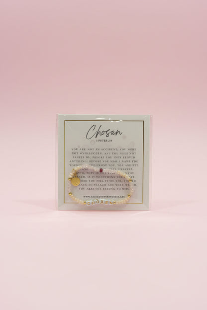 A hand holds a small gold beaded bracelet on a branded display card labeled 'Chosen Letters of Life' in a jewelry shop. The background shows pink display stands and other jewelry items in a softly blurred store setting.