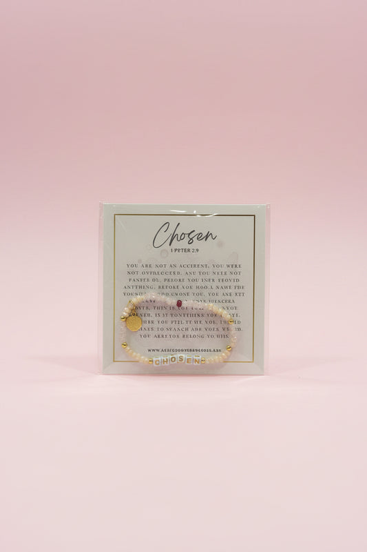 A hand holds a small gold beaded bracelet on a branded display card labeled 'Chosen Letters of Life' in a jewelry shop. The background shows pink display stands and other jewelry items in a softly blurred store setting.