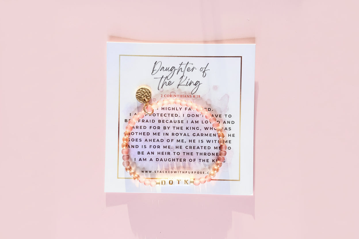 A bracelet with gold and pink beads, featuring a 'Daughter of the King' label and a quote, presented on a white card with a gold textured background.