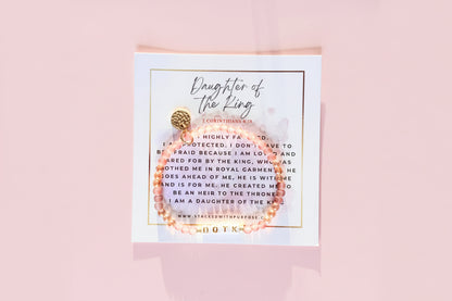 A bracelet with gold and pink beads, featuring a 'Daughter of the King' label and a quote, presented on a white card with a gold textured background.