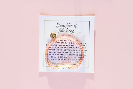 A bracelet with gold and pink beads, featuring a 'Daughter of the King' label and a quote, presented on a white card with a gold textured background.