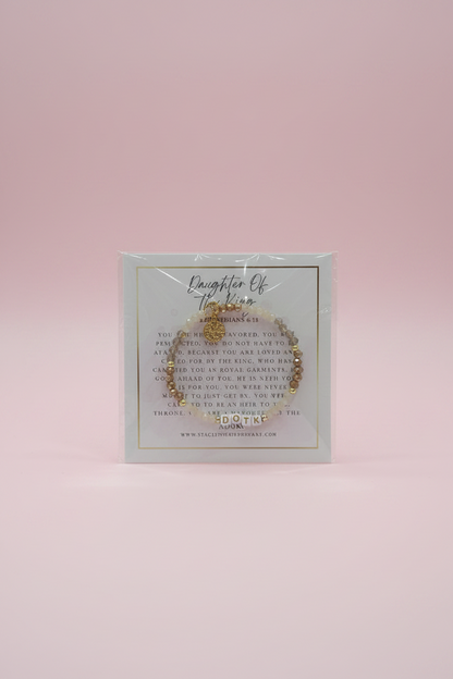 A hand holds a clear plastic card displaying a delicate gold-toned beaded bracelet with a small charm, set against a softly blurred pink jewelry display background.