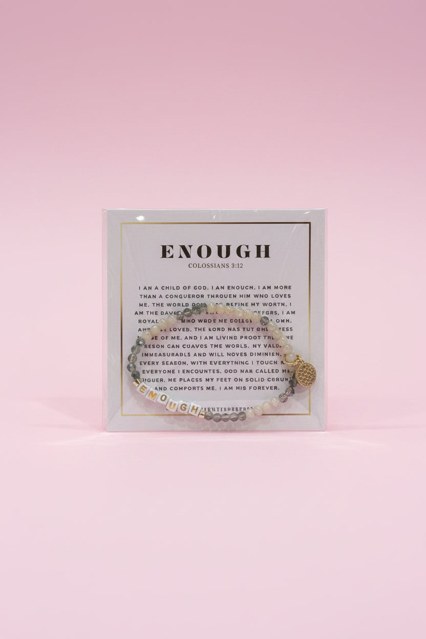 A hand holds a clear square display card featuring a silver-toned chain bracelet with a small heart charm. The card has printed text and the word 'ENOUGH' at the top.