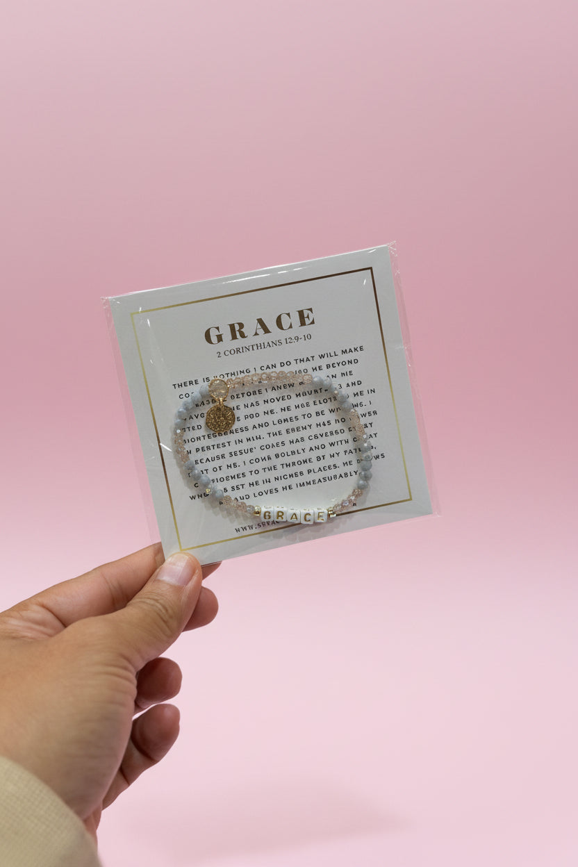 A hand holds a small square card displaying a bracelet set with a circular charm and matching beaded bracelet. The card is labeled 'GRACE' and the background shows a pink jewelry display in a store.