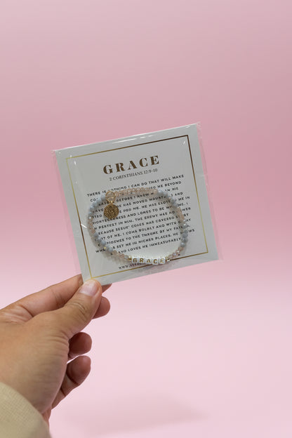 A hand holds a small square card displaying a bracelet set with a circular charm and matching beaded bracelet. The card is labeled 'GRACE' and the background shows a pink jewelry display in a store.
