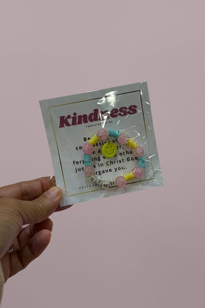 A hand holds a packaged beaded bracelet card featuring the word 'Kindness' and a small quote, with a pink jewelry display blurred in the background.