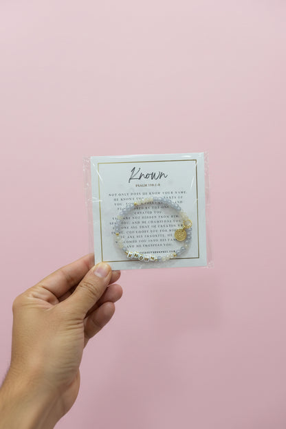 A hand holds a small jewelry card package in a shop setting. The card is white with gold border and the brand name 'Known' printed at the top, and a small gold-toned jewelry piece is attached.