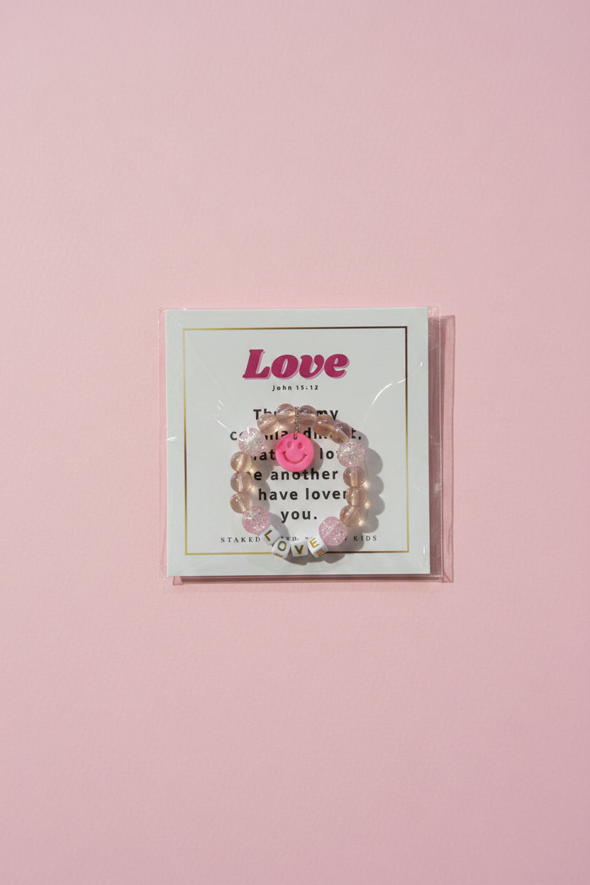 A hand holds a clear plastic-wrapped jewelry card displaying a pink beaded bracelet with a smiley face charm. The card reads 'Love' and has decorative text, with a pink boutique display and matching boxes blurred in the background.