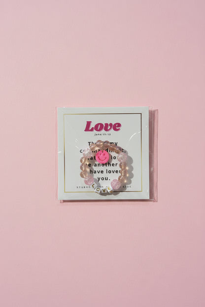 A hand holds a clear plastic-wrapped jewelry card displaying a pink beaded bracelet with a smiley face charm. The card reads 'Love' and has decorative text, with a pink boutique display and matching boxes blurred in the background.