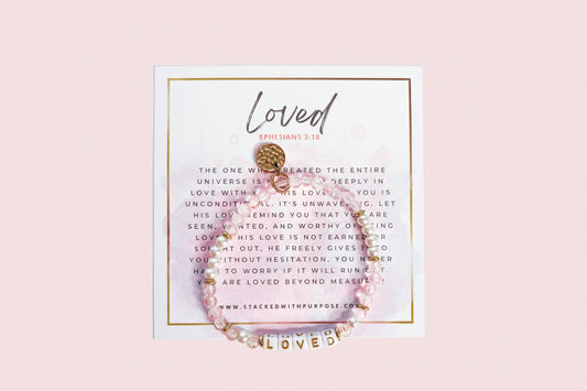 A light pink bracelet with gold accents and a gold-tone bead, featuring the word 'LOVED' and a personalized message on a stack of cards with a pink background.