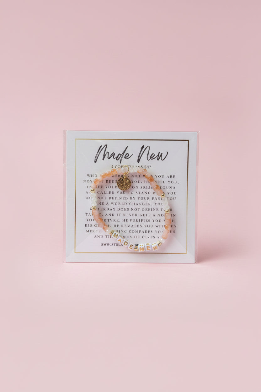 A hand holds a clear plastic card displaying a delicate gold-toned beaded bracelet with a central charm, set against a softly blurred pink jewelry store background.