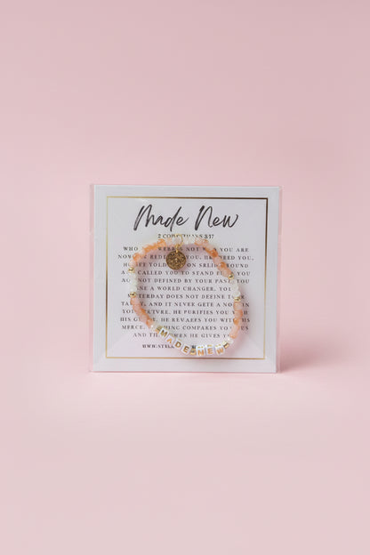 A hand holds a clear plastic card displaying a delicate gold-toned beaded bracelet with a central charm, set against a softly blurred pink jewelry store background.