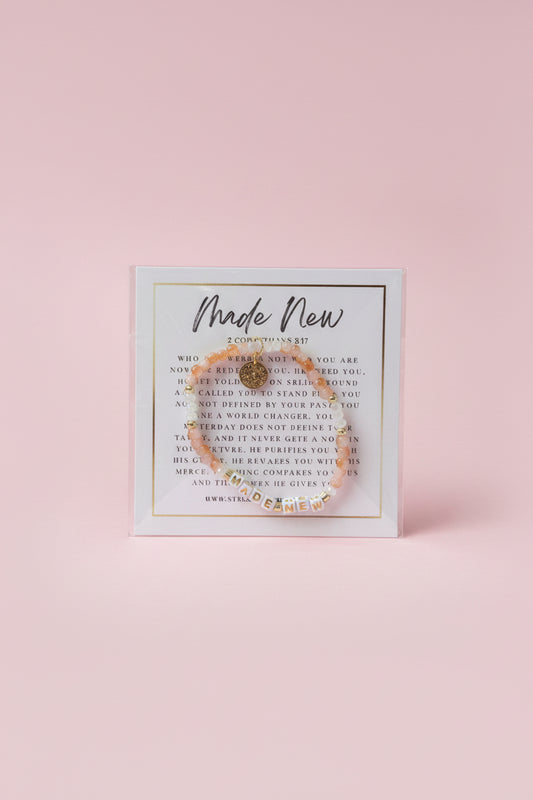 A hand holds a clear plastic card displaying a delicate gold-toned beaded bracelet with a central charm, set against a softly blurred pink jewelry store background.