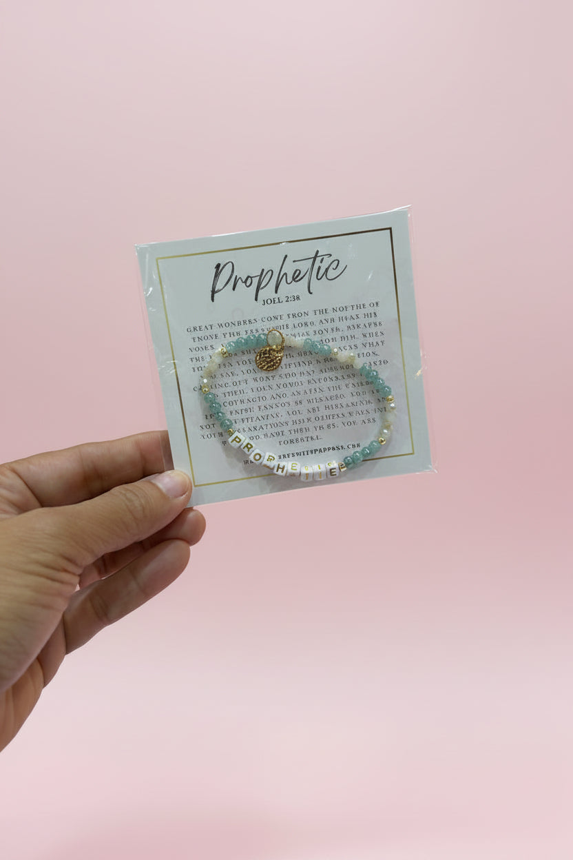 A hand holds a small jewelry card displaying a beaded bracelet with a gold heart charm. The card is branded 'Prophetic' and is photographed in a pink boutique setting with display stands in the blurred background.