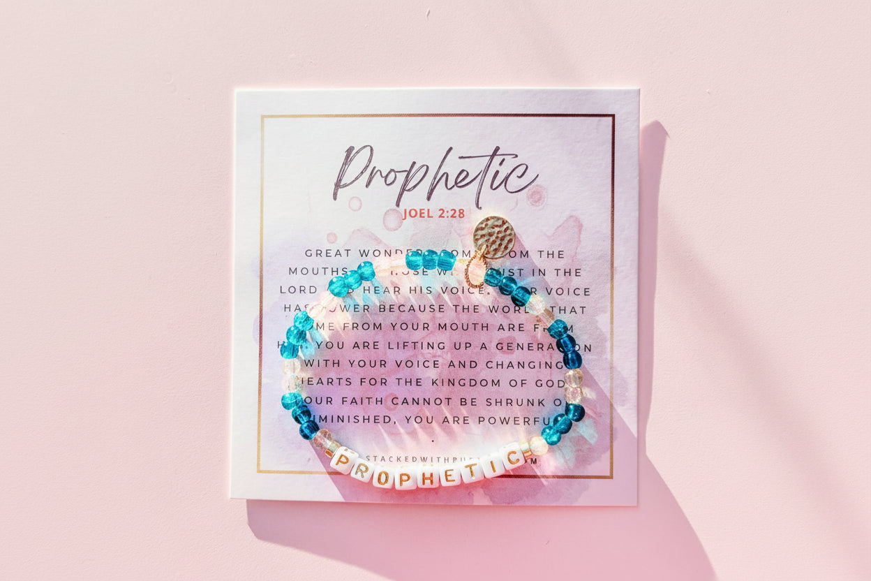 A bracelet with blue beads and a gold-colored bead, placed on a card with the word 'PROPHETIC' and a scripture verse.