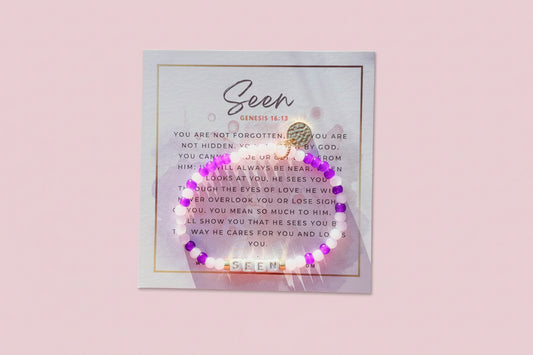 A purple bracelet with white and gold accents displayed on a card with a religious message.