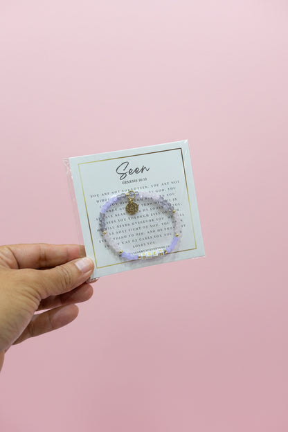 A hand holds a small bracelet on a display card in a jewelry store. The bracelet is light purple with a small gold charm and clear rhinestone accents.