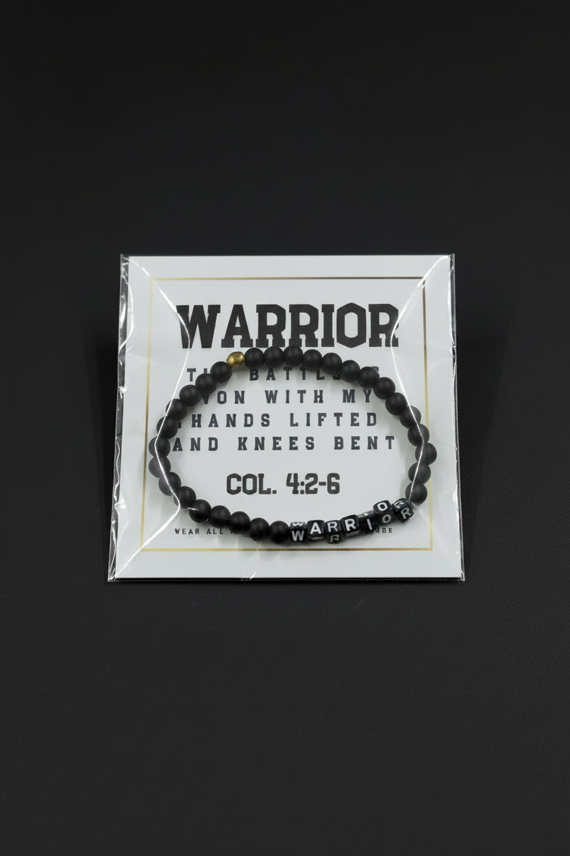 A black beaded bracelet with a silver 'WARRIOR' letter charm and a small gold accent bead, displayed on a white backing card held by a hand.