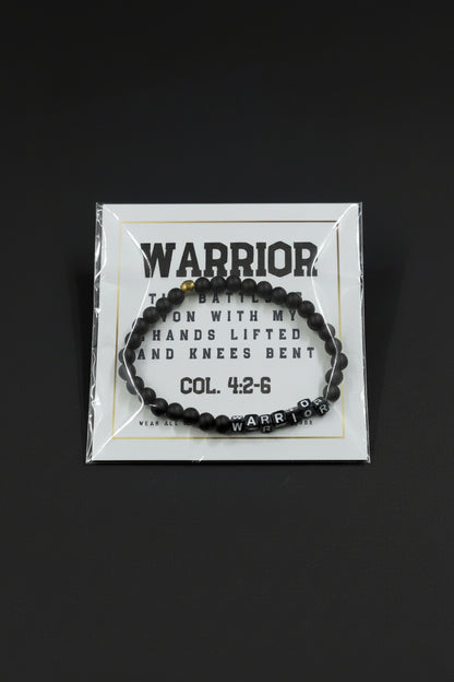 A black beaded bracelet with a silver 'WARRIOR' letter charm and a small gold accent bead, displayed on a white backing card held by a hand.
