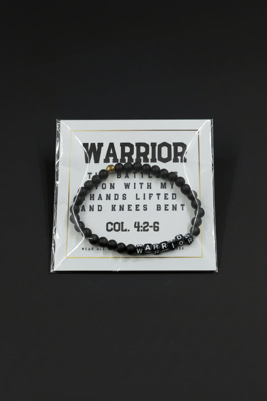 A black beaded bracelet with a silver 'WARRIOR' letter charm and a small gold accent bead, displayed on a white backing card held by a hand.