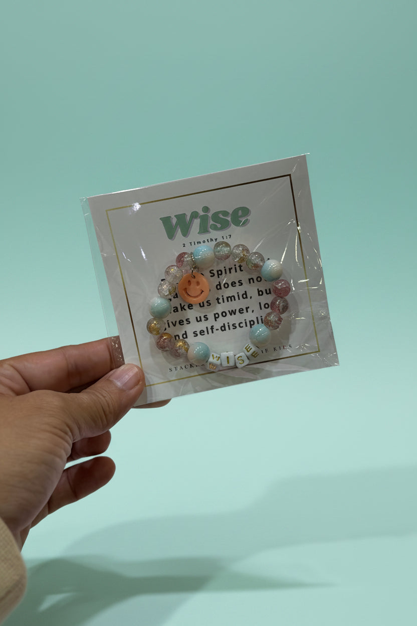 A hand holds a packaged beaded bracelet with a smiley face charm and letter beads spelling 'WISDOM' against a pink jewelry display background.