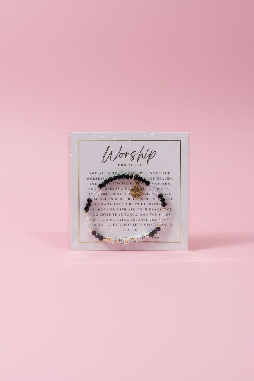 A hand holds a small beaded bracelet on a printed display card in a jewelry store. The bracelet has dark beads, a small gold-toned central charm, and tiny clear rhinestone accents.