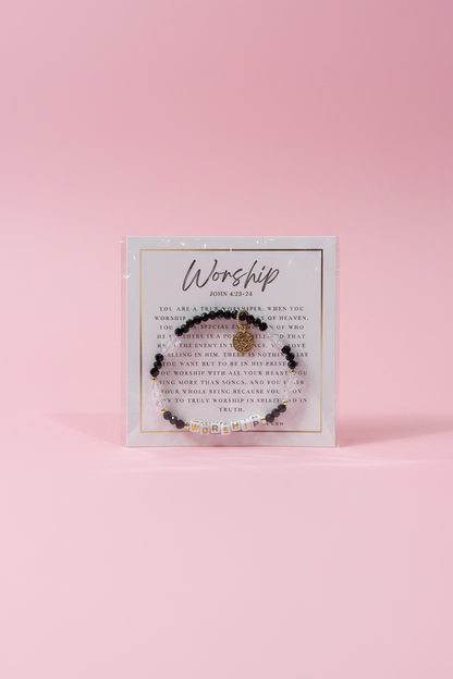 A hand holds a small beaded bracelet on a printed display card in a jewelry store. The bracelet has dark beads, a small gold-toned central charm, and tiny clear rhinestone accents.
