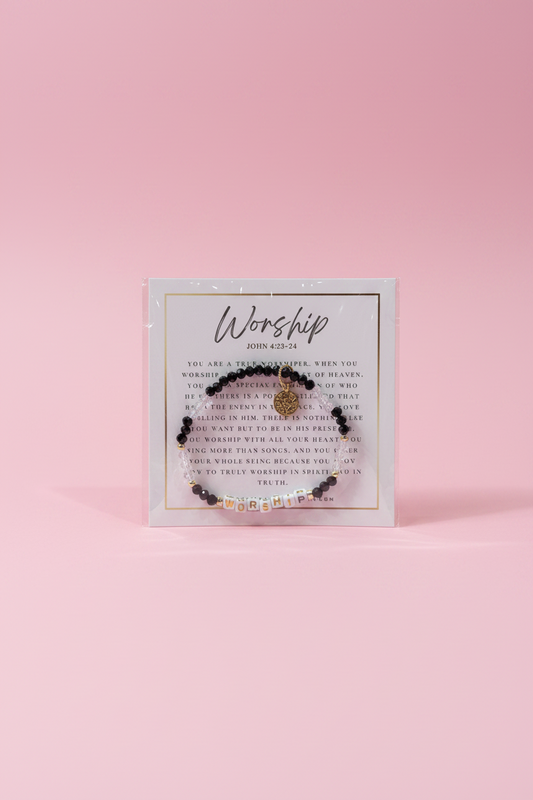 A hand holds a small beaded bracelet on a printed display card in a jewelry store. The bracelet has dark beads, a small gold-toned central charm, and tiny clear rhinestone accents.