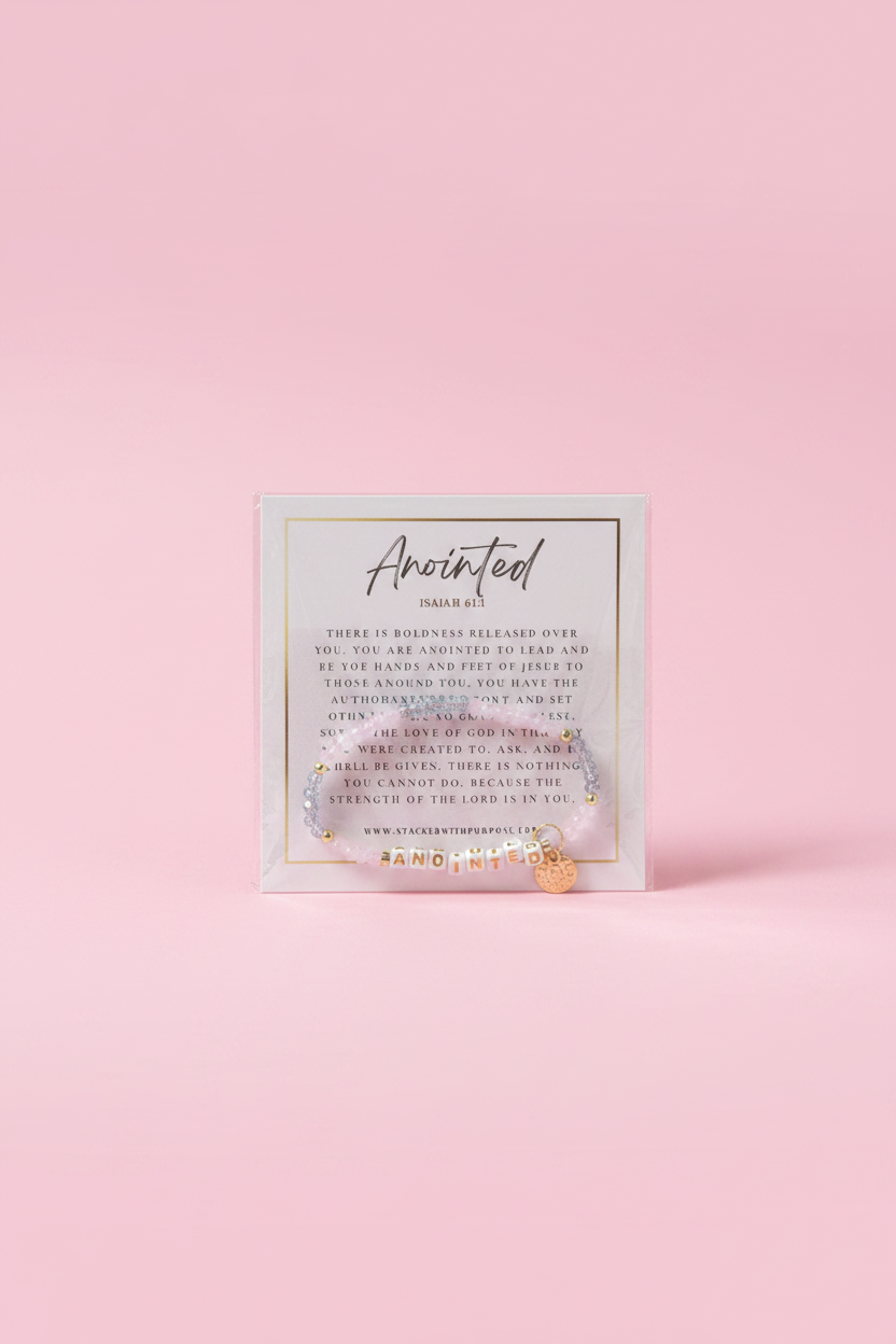 A hand holding a small square card with the word 'Anointed' and inspirational text, set against a softly blurred pink boutique background.