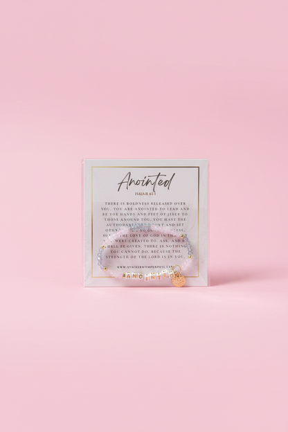 A hand holding a small square card with the word 'Anointed' and inspirational text, set against a softly blurred pink boutique background.