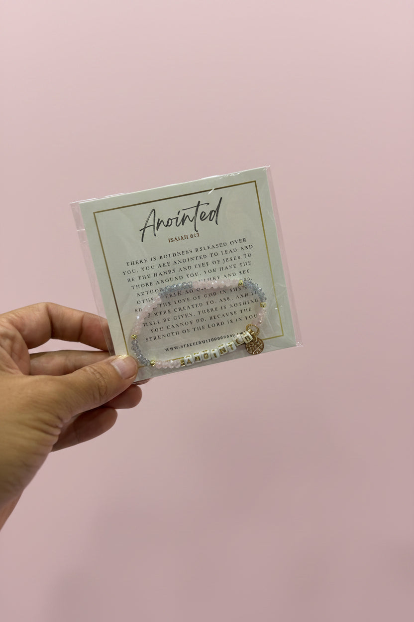 A hand holds a clear plastic sleeve containing a square card with the phrase 'Aloved Again' in script and small gold-toned jewelry attached at the bottom, set against a blurred pink jewelry display.