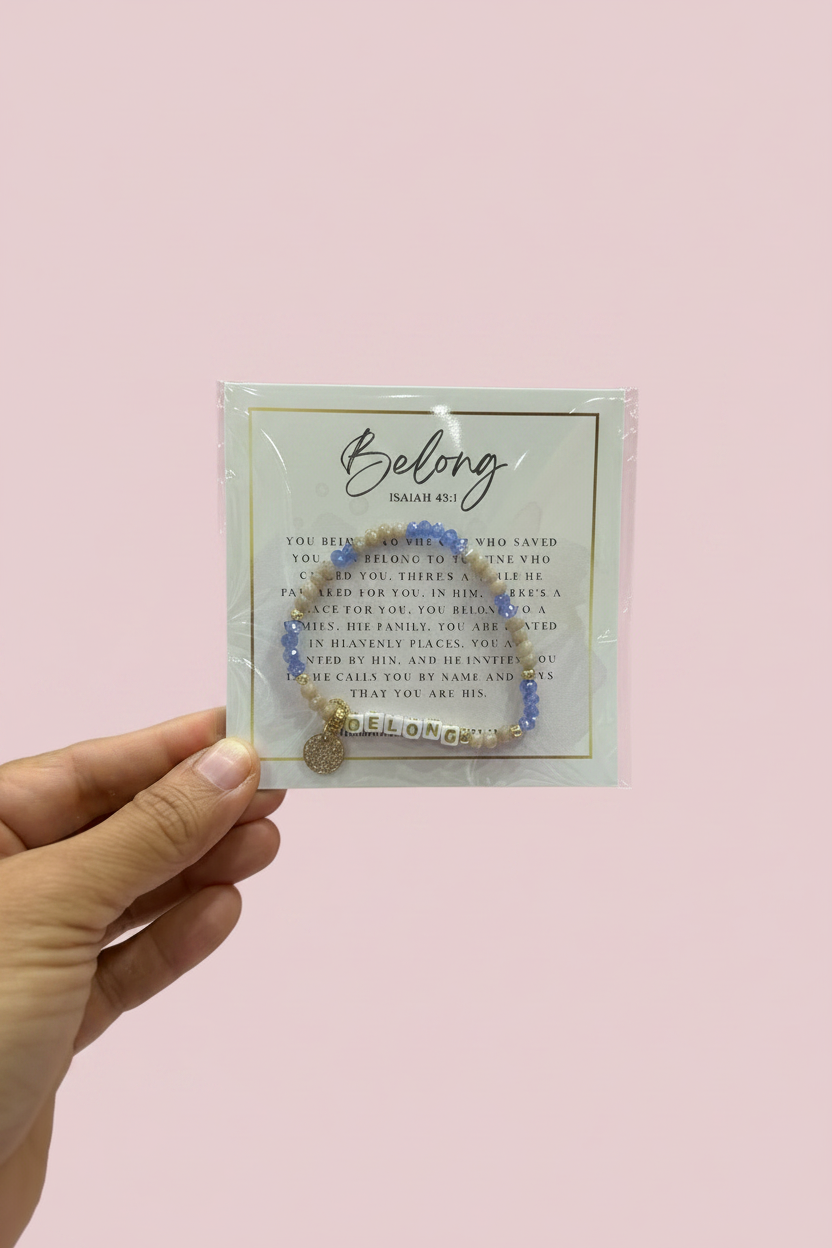 A hand holding a packaged beaded bracelet against a solid light pink background. The bracelet features light blue and white beads with a gold-toned metal charm and a gold heart, displayed on a white backing card with the brand name 'Belong Online US'.