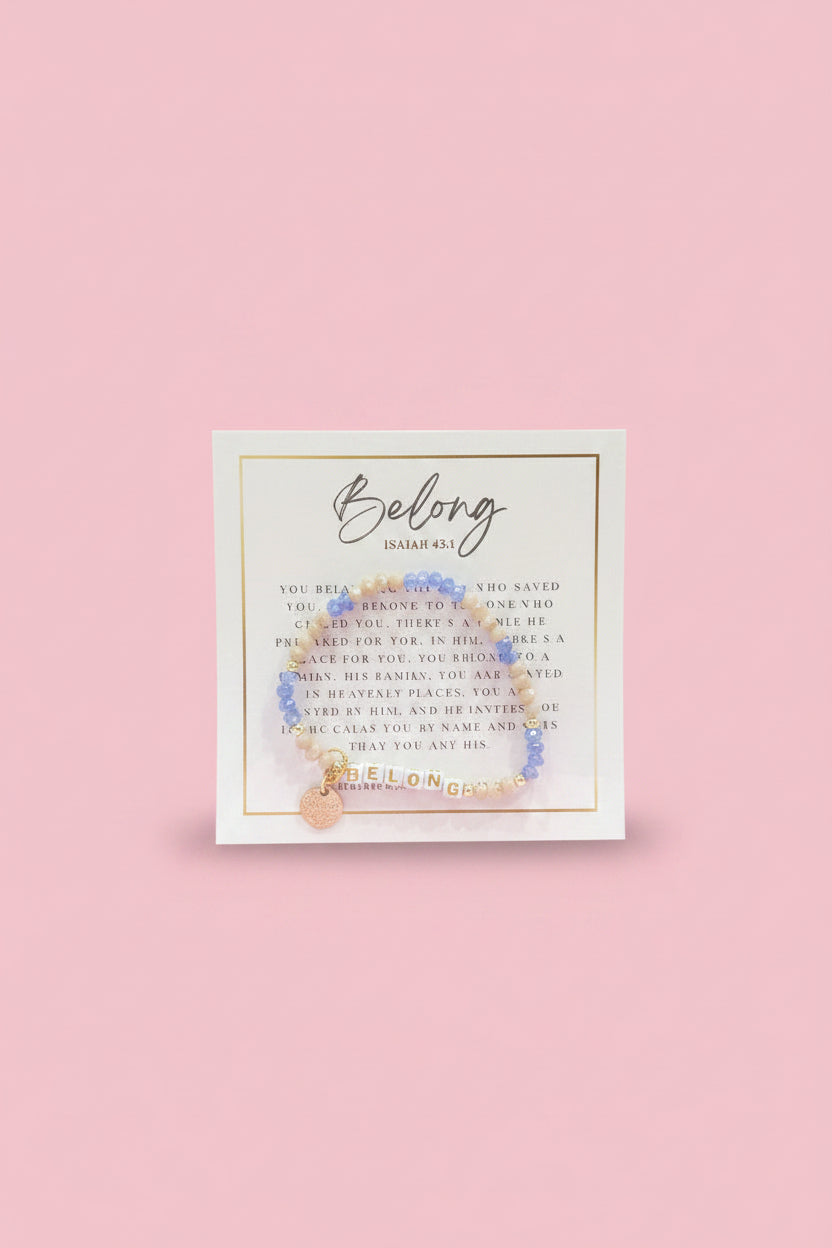 A hand holds a small beaded bracelet on a branded display card in a shop setting. The bracelet has light blue beads, a gold-toned connector, and a small gold charm.