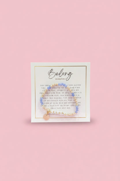 A hand holds a small beaded bracelet on a branded display card in a shop setting. The bracelet has light blue beads, a gold-toned connector, and a small gold charm.