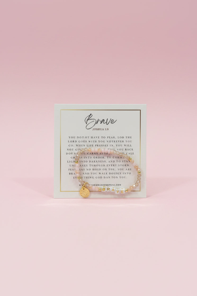A hand holds a small gold beaded bracelet on a printed display card in a jewelry store. The card reads 'Brave' and 'Godspeed,' and the background shows pink display boxes and other jewelry items.