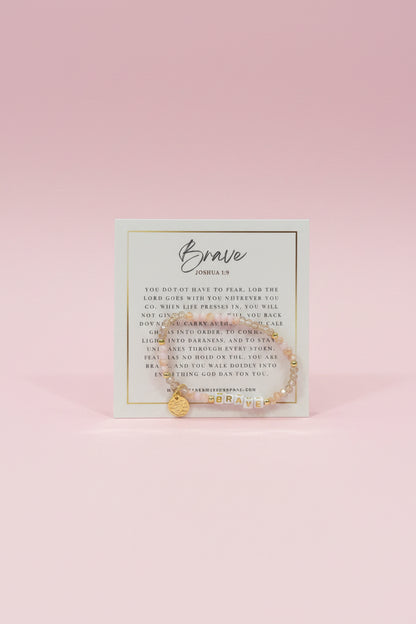 A hand holds a small gold beaded bracelet on a printed display card in a jewelry store. The card reads 'Brave' and 'Godspeed,' and the background shows pink display boxes and other jewelry items.