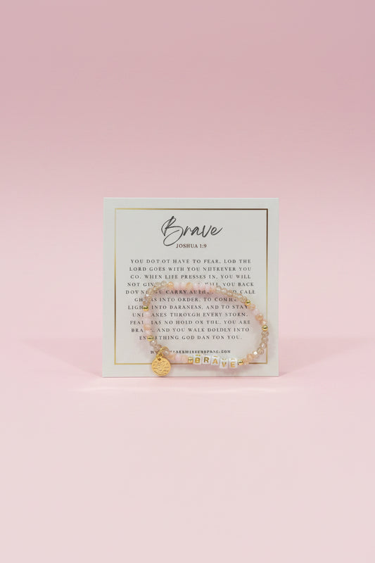 A hand holds a small gold beaded bracelet on a printed display card in a jewelry store. The card reads 'Brave' and 'Godspeed,' and the background shows pink display boxes and other jewelry items.