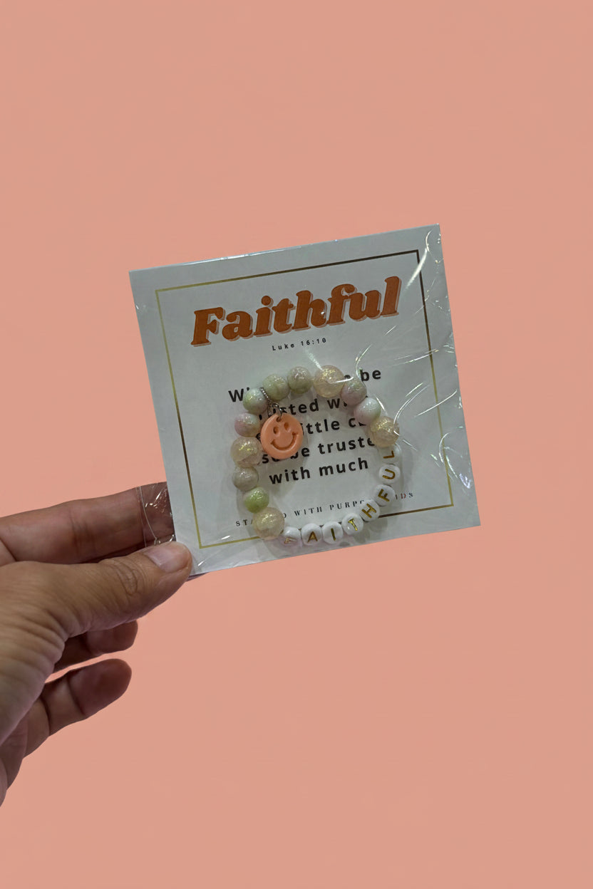 A hand holds a packaged beaded bracelet on a display card labeled 'Faithful' in a jewelry store. The bracelet has cream-colored beads with a small peach smiley face bead and white letter beads spelling 'FAITHFUL'.