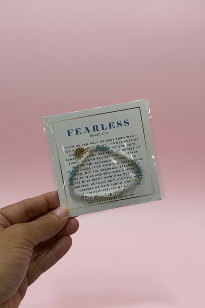 A hand holds a small jewelry card displaying a silver-toned bracelet with a gold-toned charm, set against a softly blurred pink retail display background.