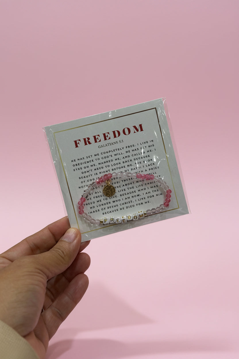 A hand holds a small jewelry card displaying a pink beaded bracelet with a gold heart charm and a white beaded bracelet. The card is labeled 'FREEDOM' and is photographed in a pink boutique setting with blurred display stands in the background.