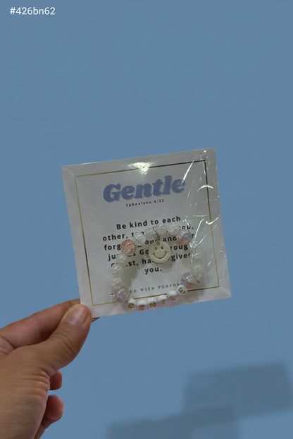 A hand holds a clear plastic-wrapped jewelry card featuring a small beaded bracelet with a smiley face charm and pearl-like beads. The card reads 'Gentle' and has a sentimental quote.
