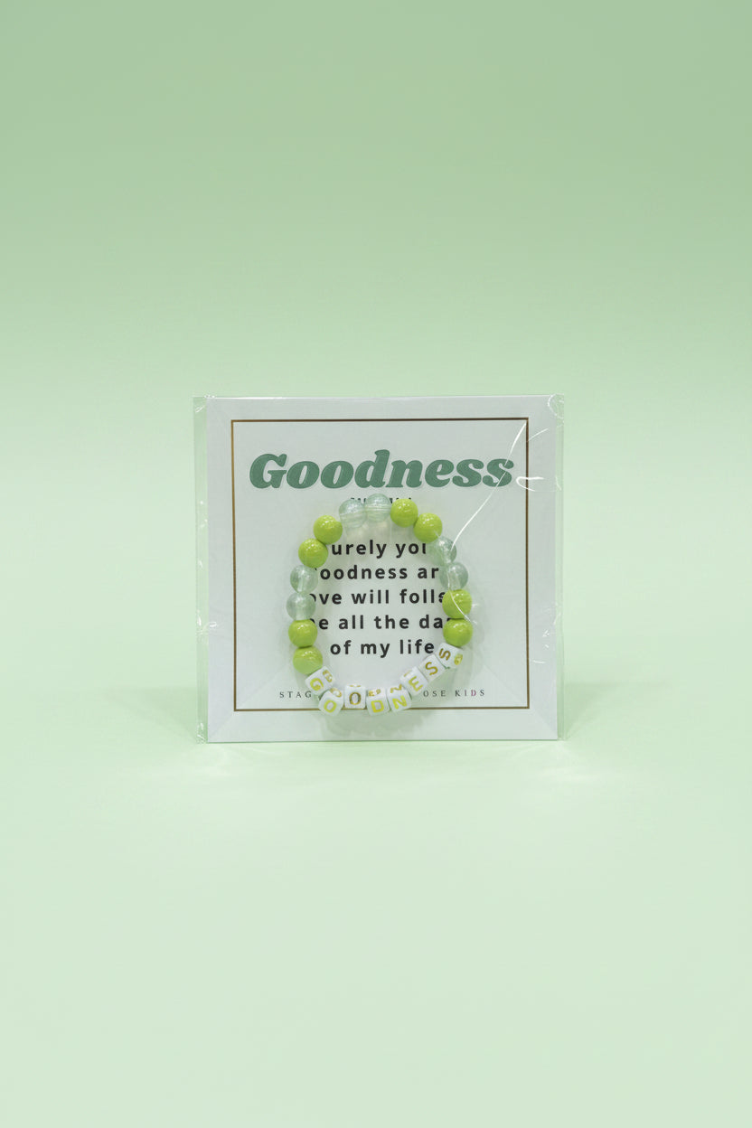 A hand holds a packaged beaded bracelet on a printed card that reads 'Goodness' in green text, with a pink and cream boutique display blurred in the background.